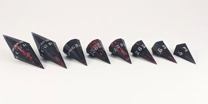 A straight on photo of a complete set of dagger DnD dice that are black with crimson red splatters with silver numbers arranged in a straight line with points angled toward the right. They are lined up with the 2 d20s on the left going down to the d4 on the right side. The highest value number of each die is facing forward.