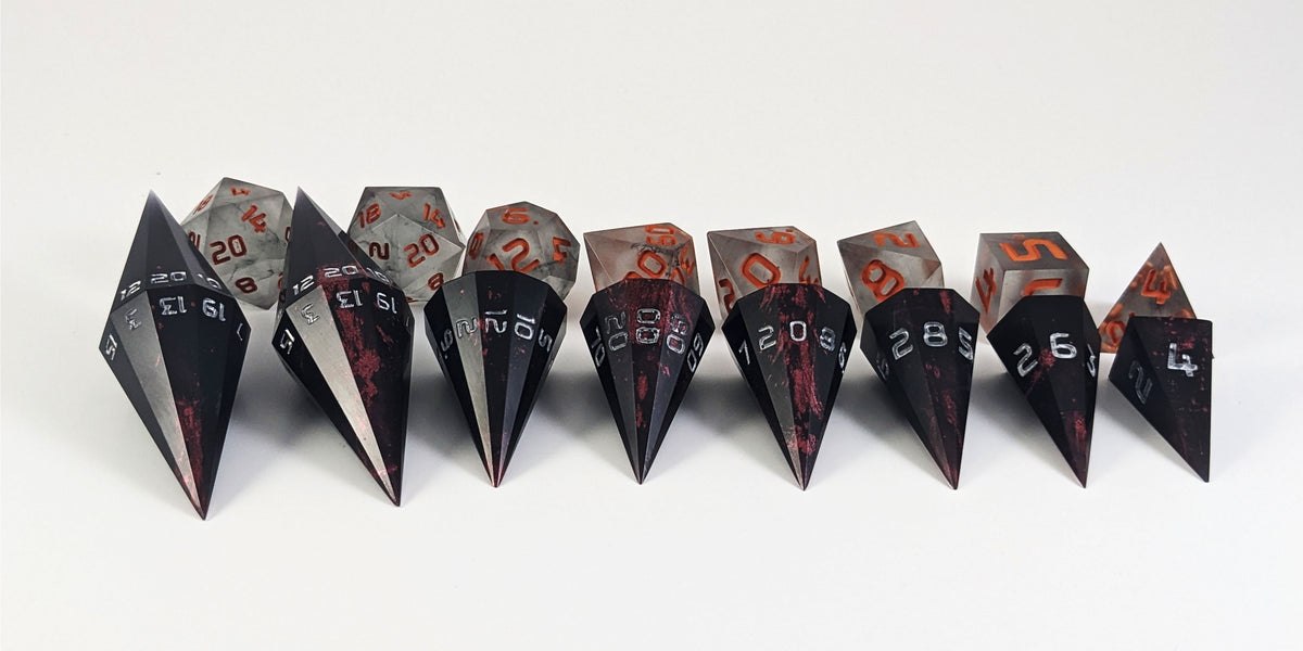 A straight on photo of a complete set of dagger dice that are black with crimson red splatters with silver numbers arranged in a straight line with points angled forward. They are lined up with the 2 d20s on the left going down to the d4 on the right side. Behind the dagger set is a standard set of polyhedral dnd dice to allow for size comparison. The dagger dice are much larger overall, but not taller. The highest value number of each dagger die is facing up.