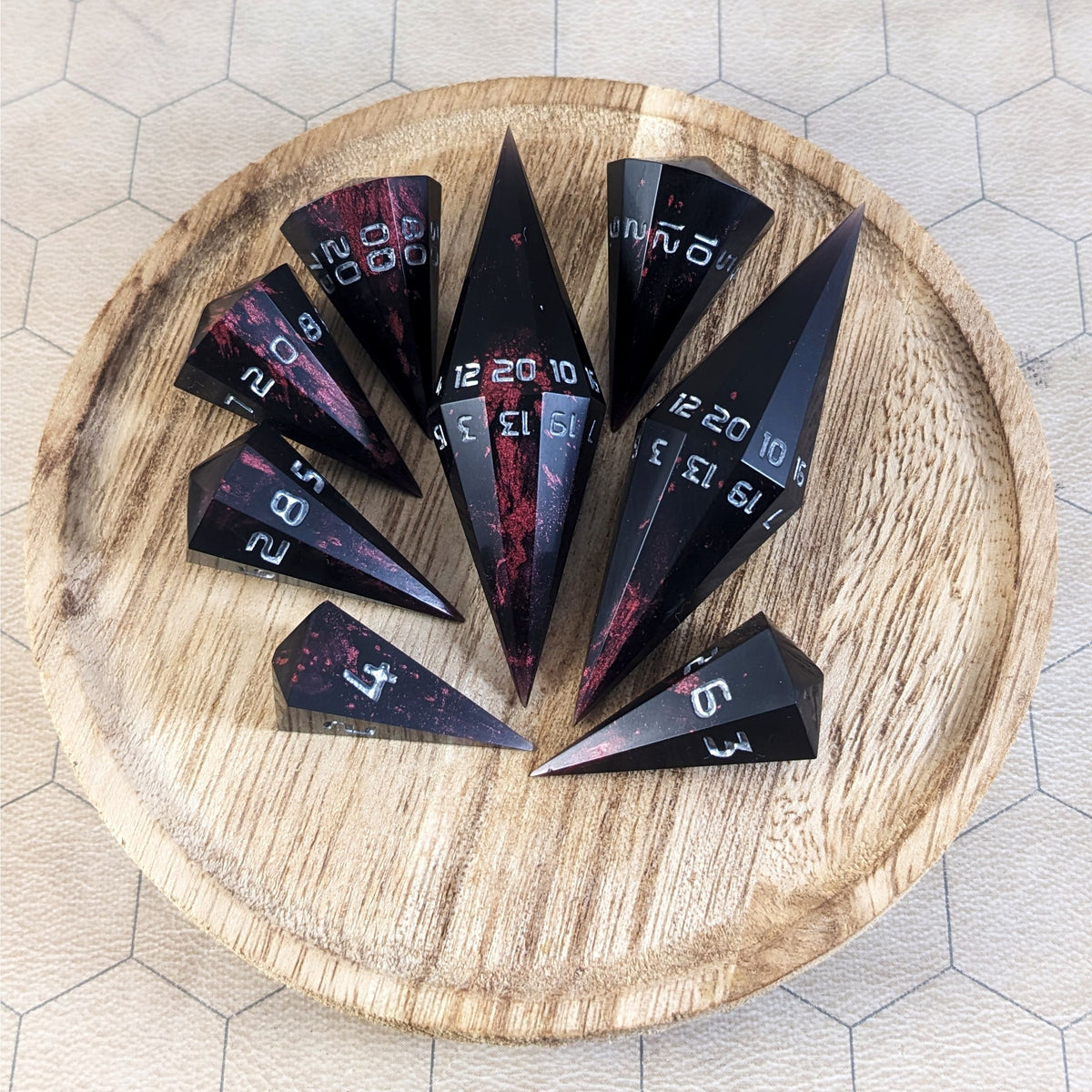 A complete set of dagger style resin dice that are black with crimson red marbling and silver numbers are arranged on a wooden circle. The 2 d20s are next to each other with the d12 placed between them on top. The d6 is to the bottom right of the d20s and the rest of the dice are arranged along the left side, points angled toward the d20s.