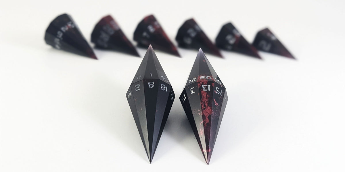 A straight on photo of a dagger set of DnD dice that are black with splatters of crimson red with silver numbers focusing on 2 d20s in the center. The d20 on the left has the 1 face up and the d20 on the right has the 20 face up. The other 6 dice are lined up in the background but are out of focus, while the d20s are in sharp focus.