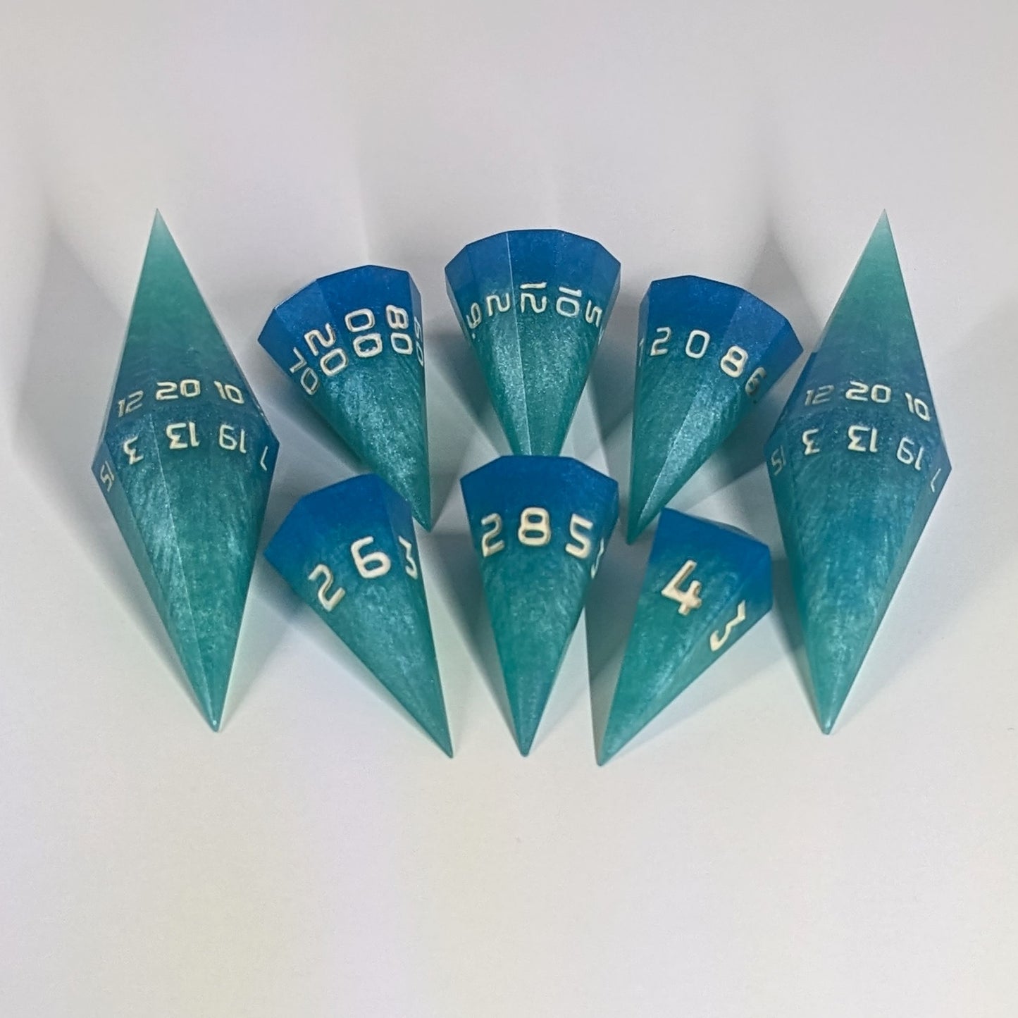 A dagger style DnD dice set that is  oceanic shades of blue and green with sandy white numbers is arranged with a d20 each on the right and left, with the d%, d12, and d10 along the top and the d6, d8, and d4 along the bottom, all in line with the d20s.
