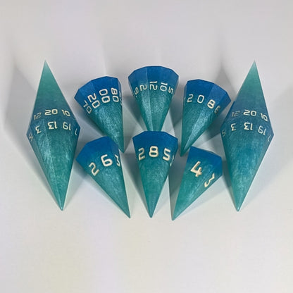 A dagger style DnD dice set that is  oceanic shades of blue and green with sandy white numbers is arranged with a d20 each on the right and left, with the d%, d12, and d10 along the top and the d6, d8, and d4 along the bottom, all in line with the d20s.