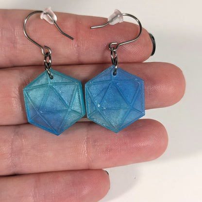 A pair of oceanic blue and green dangly, flat d20 earrings with silver color titanium and niobium hardware with bell shaped rubber backs held on on a hand on a white background.