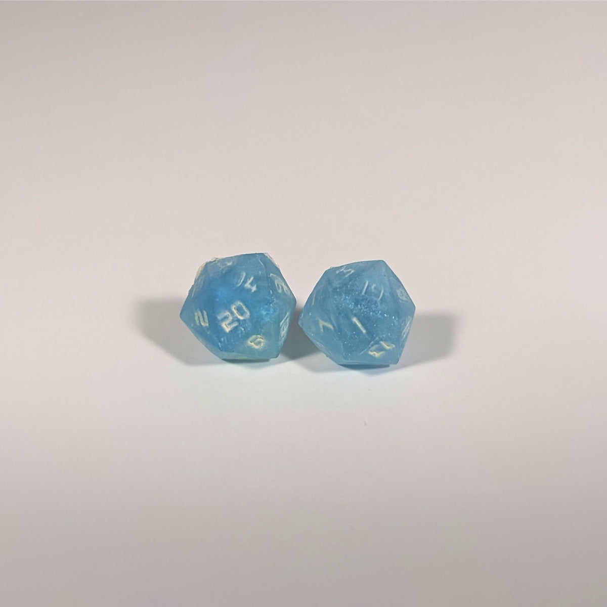 A single pair of the larger size handmade d20 earrings with titanium hardware that are oceanic shades of blue and green with sandy white numbers on a white background. The earring on the left centers the 20 face and the earring on the right centers the 1 face.