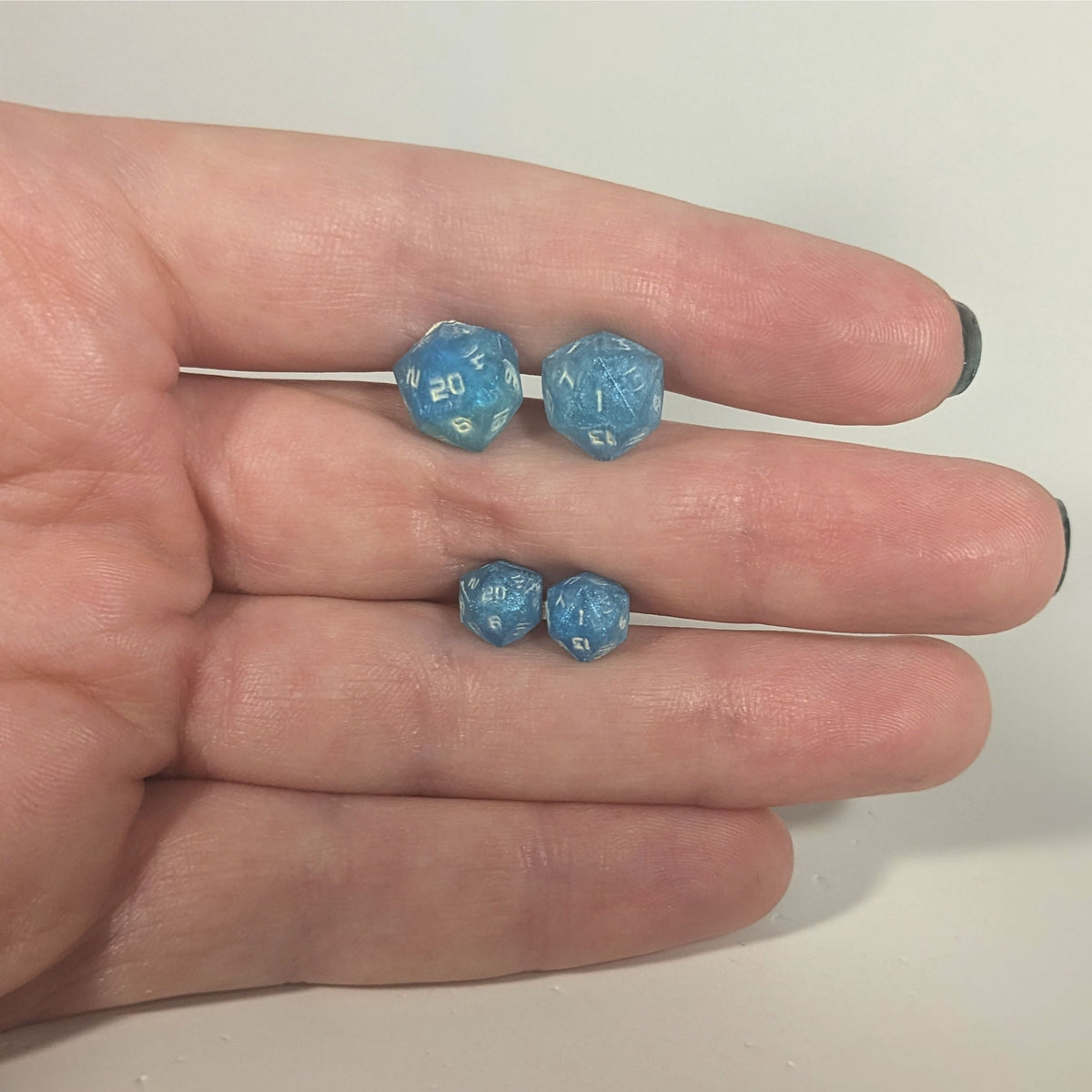 Two pairs of handmade d20 earrings with titanium hardware that are oceanic shades of blue and green with sandy white numbers held between the fingers of a flat hand. The larger pair is between the pointer and middle finger, the smaller between the middle and ring finger. The earrings on the left of each pair centers the 20 face and the earrings on the right of each pair centers the 1 face.