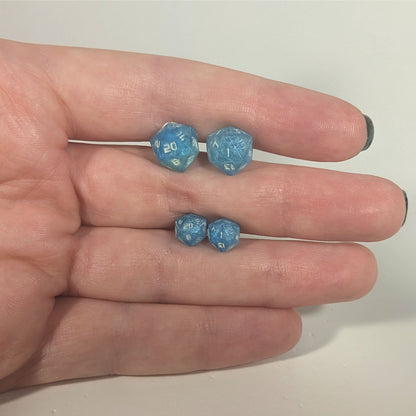 Two pairs of handmade d20 earrings with titanium hardware that are oceanic shades of blue and green with sandy white numbers held between the fingers of a flat hand. The larger pair is between the pointer and middle finger, the smaller between the middle and ring finger. The earrings on the left of each pair centers the 20 face and the earrings on the right of each pair centers the 1 face.