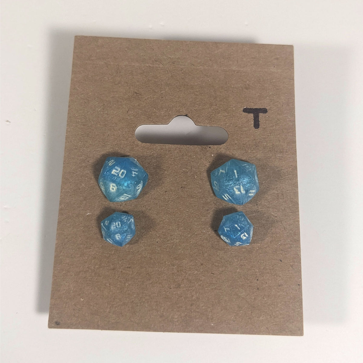 Two pairs of handmade d20 earrings with titanium hardware that are oceanic shades of blue and green with sandy white numbers on a kraft brown earring card with a T in the top right corner. The earrings on the left of each pair centers the 20 face and the earrings on the right of each pair centers the 1 face.
