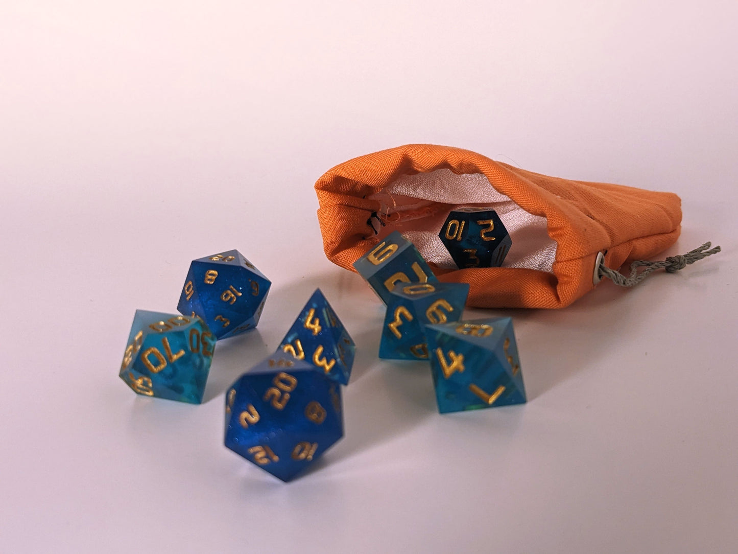 Small handmade dice bag that is orange lays flat with the top open and blue dnd dice spilling out from it.