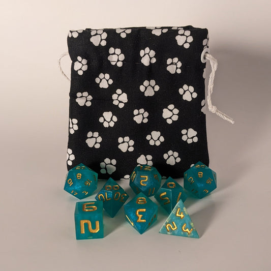 Small handmade dice bag that is black with a white paw print pattern sits upright with teal dnd dice sitting in front of it.