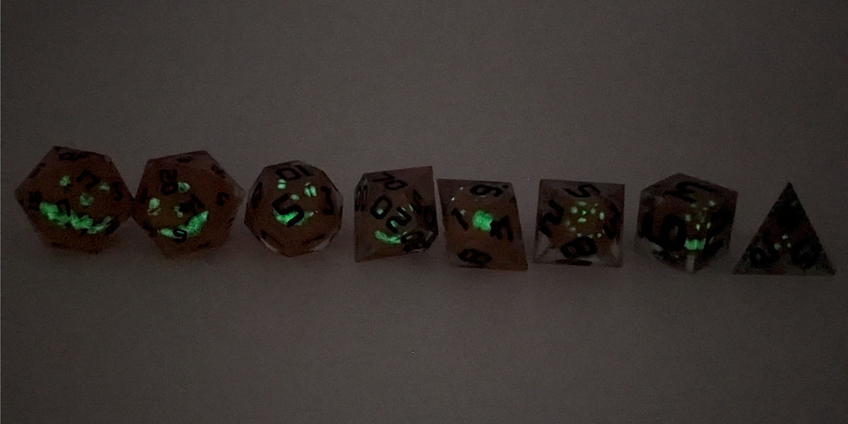 A straight on photo of a complete set of polyhedral dice that have jack-o-lantern inserts whose faces glow in the dark with dark green numbers arranged in a straight line. They are lined up with the 2 d20s on the left going down to the d4 on the right side. The dice are angled so the jack-o-lantern faces face the camera. This photo is taken in the dark to show the jack-o-lantern faces glowing green in the dark.