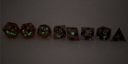 A straight on photo of a complete set of polyhedral dice that have jack-o-lantern inserts whose faces glow in the dark with dark green numbers arranged in a straight line. They are lined up with the 2 d20s on the left going down to the d4 on the right side. The dice are angled so the jack-o-lantern faces face the camera. This photo is taken in the dark to show the jack-o-lantern faces glowing green in the dark.