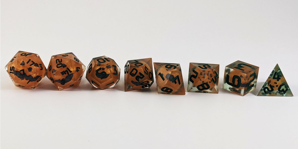 A straight on photo of a complete set of polyhedral dice that have jack-o-lantern inserts whose faces glow in the dark with dark green numbers arranged in a straight line. They are lined up with the 2 d20s on the left going down to the d4 on the right side. The dice are angled so the jack-o-lantern faces face the camera.