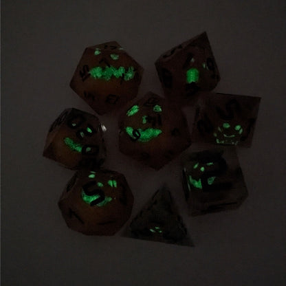 A complete set of 8 handmade artisan dice that have jack-o-lantern inserts whose faces glow in the dark with dark green numbers. The DnD dice set is arranged in a circle and features 2 D20s, one of which is in the center surrounded by the other 7 dice on a white background. The dice are arranged so the jack-o-lantern faces are facing as upwards as possible. The photo is taken in the dark to show the faces glowing green in the dark.