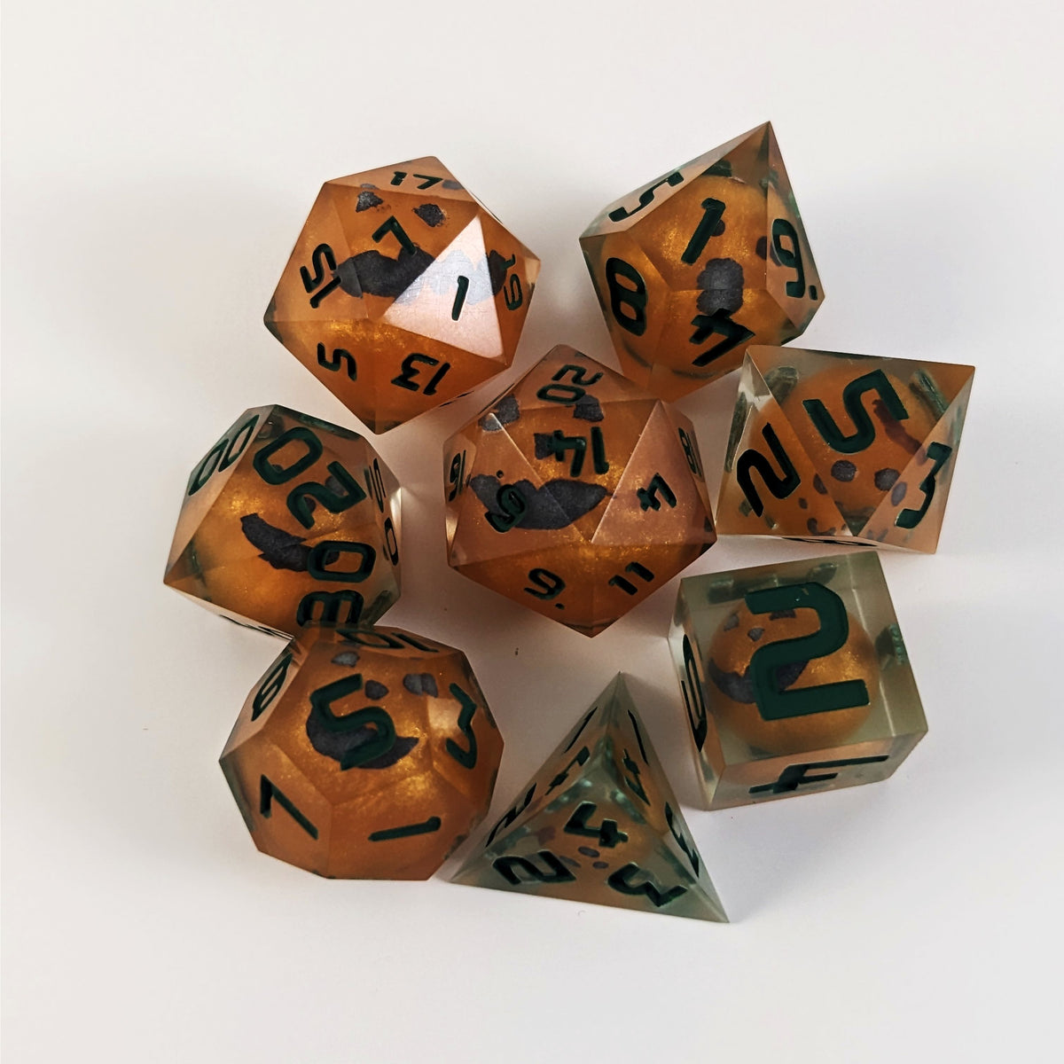 A complete set of 8 handmade artisan dice that have jack-o-lantern inserts whose faces glow in the dark with dark green numbers. The DnD dice set is arranged in a circle and features 2 D20s, one of which is in the center surrounded by the other 7 dice on a white background. The dice are arranged so the jack-o-lantern faces are facing as upwards as possible.