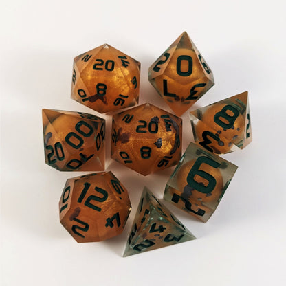 A complete set of 8 handmade artisan dice that have jack-o-lantern inserts whose faces glow in the dark with dark green numbers. The DnD dice set is arranged in a circle and features 2 D20s, one of which is in the center surrounded by the other 7 dice on a white background.