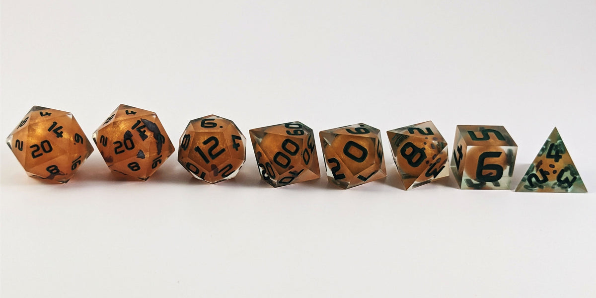 A straight on photo of a complete set of polyhedral dice that have jack-o-lantern inserts whose faces glow in the dark with dark green numbers arranged in a straight line. They are lined up with the 2 d20s on the left going down to the d4 on the right side. The highest value number of each die is facing forward