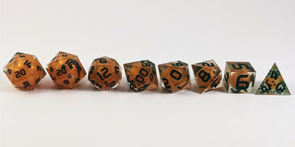 A straight on photo of a complete set of polyhedral dice that have jack-o-lantern inserts whose faces glow in the dark with dark green numbers arranged in a straight line. They are lined up with the 2 d20s on the left going down to the d4 on the right side. The highest value number of each die is facing forward