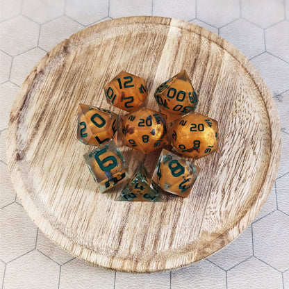 A complete set of 8 handmade artisan dice that have jack-o-lantern inserts whose faces glow in the dark with dark green numbers. The DnD dice set is on a wooded circle and the dice are arranged in a circle and features 2 D20s, one of which is in the center surrounded by the other 7 dice.