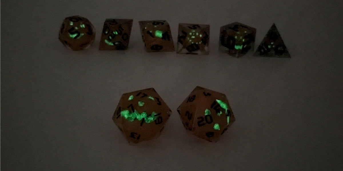 A straight on photo of a standard set of DnD dice that have jack-o-lantern inserts whose faces glow in the dark with dark green numbers focusing on 2 d20s in the center. The d20 on the left has the 1 face centered and the d20 on the right has the 20 face centered. The other 6 dice are lined up in the background but are out of focus, while the d20s are in sharp focus. This photo is taken in the dark to show the jack-o-lantern faces glowing green in the dark.