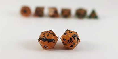 A straight on photo of a standard set of DnD dice that are have jack-o-lantern inserts whose faces glow in the dark with dark green numbers focusing on 2 d20s in the center. The d20 on the left has the 1 face centered and the d20 on the right has the 20 face centered. The other 6 dice are lined up in the background but are out of focus, while the d20s are in sharp focus.