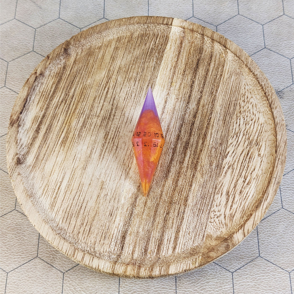 An orange and purple diamond d20 with black numbers sits in the center of a wooden circle on a beige hex map.