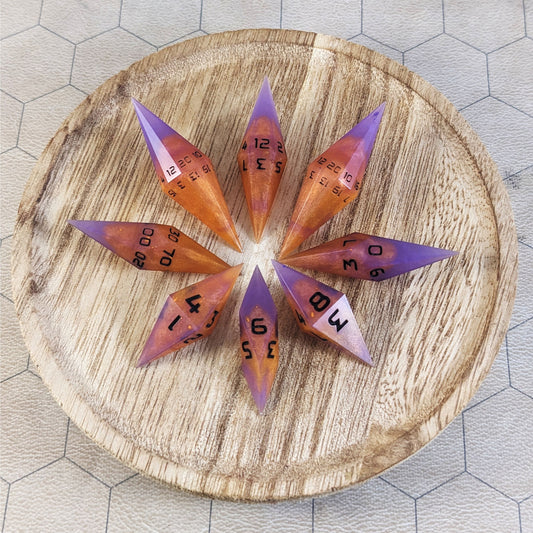 A set of handmade diamond dice that are purple and orange with black numbers arranged on a wooden circle. The resin dice have their tips facing outward, and the highest number of each die is facing up.