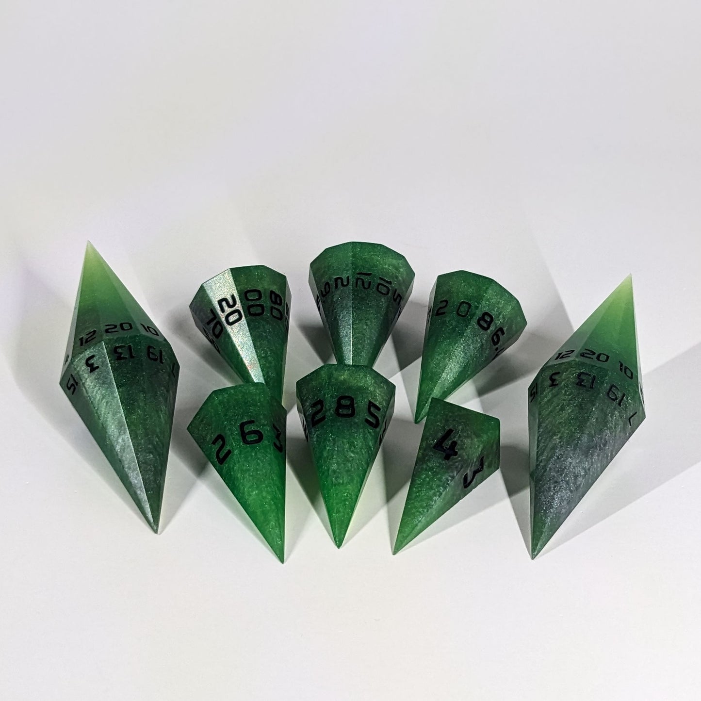 A dagger style DnD dice set that is venomous green with hints of silver like a poisoned blade with black numbers is arranged with a d20 each on the right and left, with the d%, d12, and d10 along the top and the d6, d8, and d4 along the bottom, all in line with the d20s.