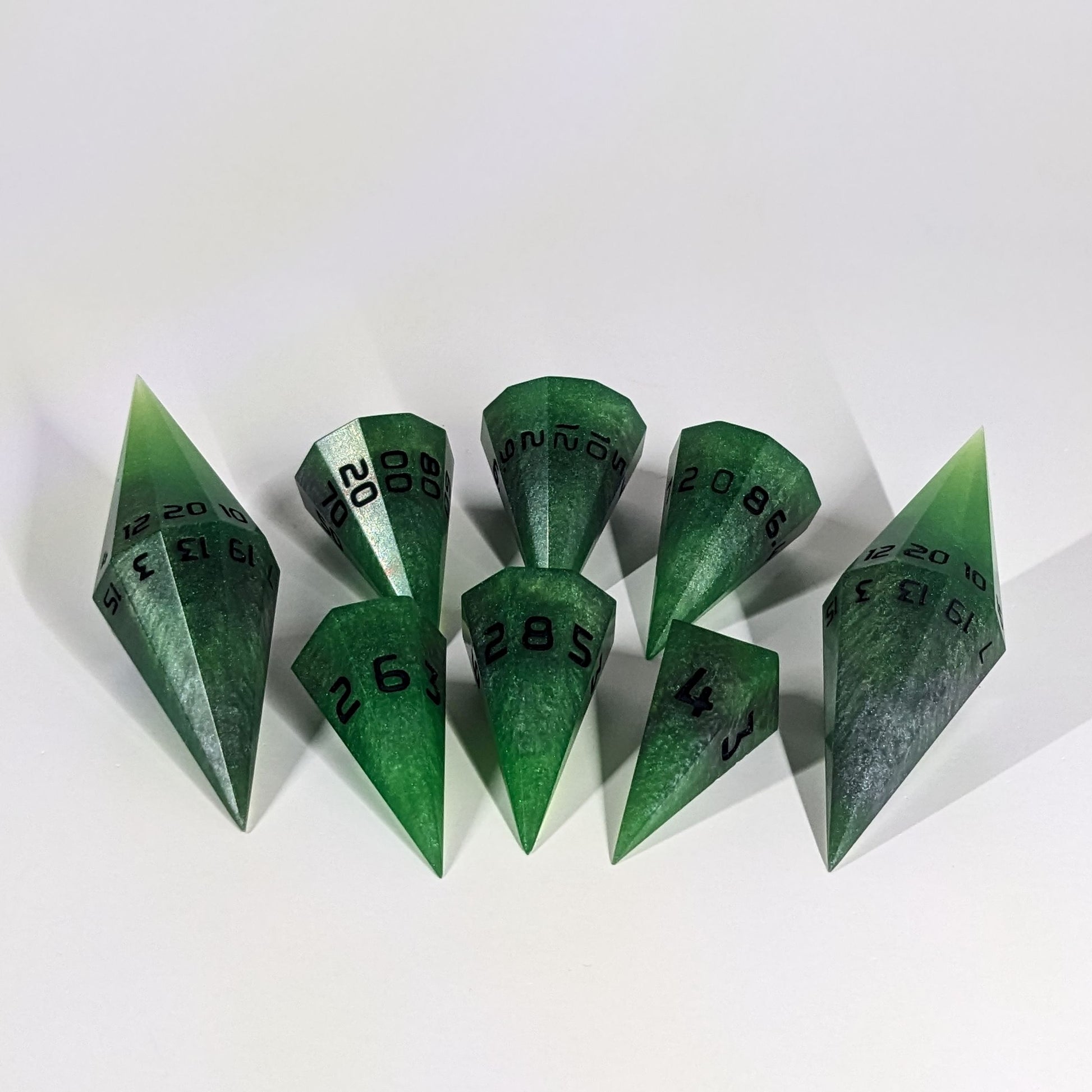 A dagger style DnD dice set that is venomous green with hints of silver like a poisoned blade with black numbers is arranged with a d20 each on the right and left, with the d%, d12, and d10 along the top and the d6, d8, and d4 along the bottom, all in line with the d20s.