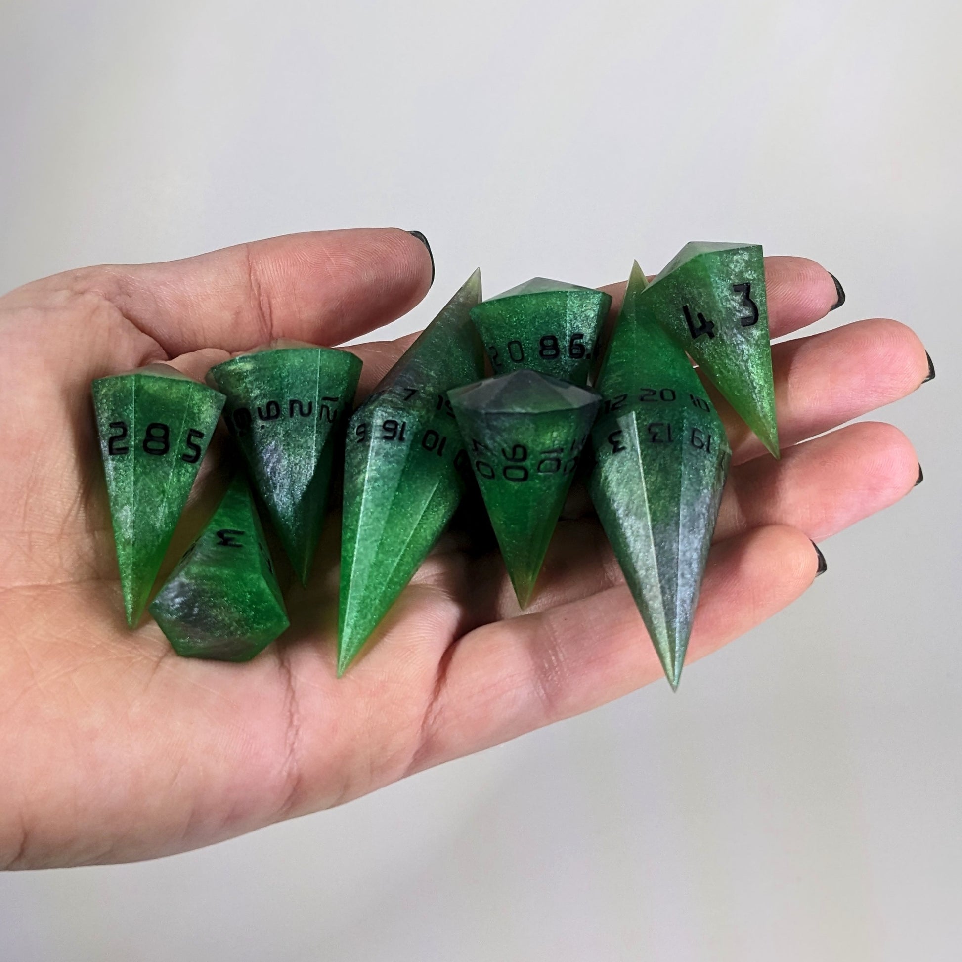 An 8 dice set of dagger style dice that are venomous green with hints of silver like a poisoned blade with black numbers are held in a hand. The image shows that the set just fits in a hand, making them overall larger than a standard set of dnd dice.