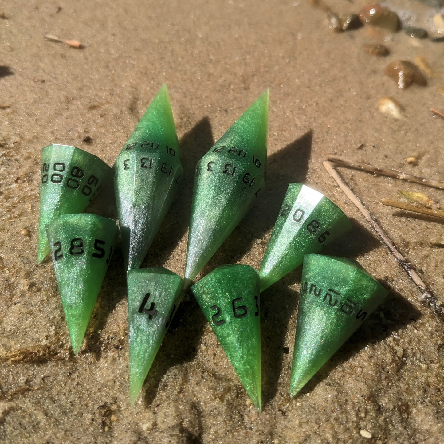 A dagger shaped dnd dice set that is venomous green with hints of silver like a poisoned blade with black numbers sits on a sandy beach in direct sunlight with a small glimpse of water in the upper right hand corner.