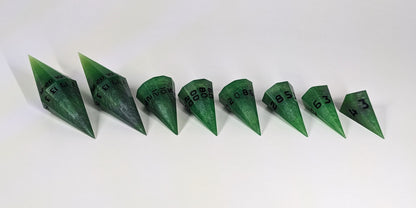 A straight on photo of a complete set of dagger DnD dice that are venomous green with hints of silver like a poisoned blade with black numbers arranged in a straight line with points angled toward the right. They are lined up with the 2 d20s on the left going down to the d4 on the right side. The highest value number of each die is facing forward.