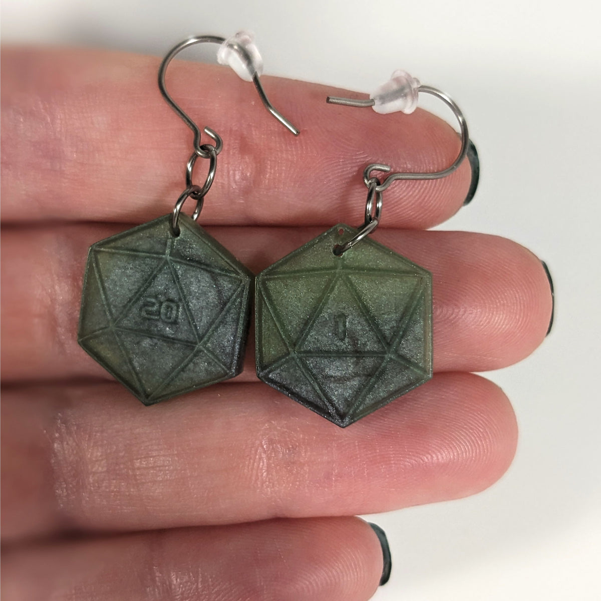 A pair of dark green and dark silver dangly, flat d20 earrings with silver color titanium and niobium hardware with bell shaped rubber backs held on on a hand on a white background. The earring on the left has a 20 on it and the earring on the right has a 1 on it, both in a sci-fi style font.
