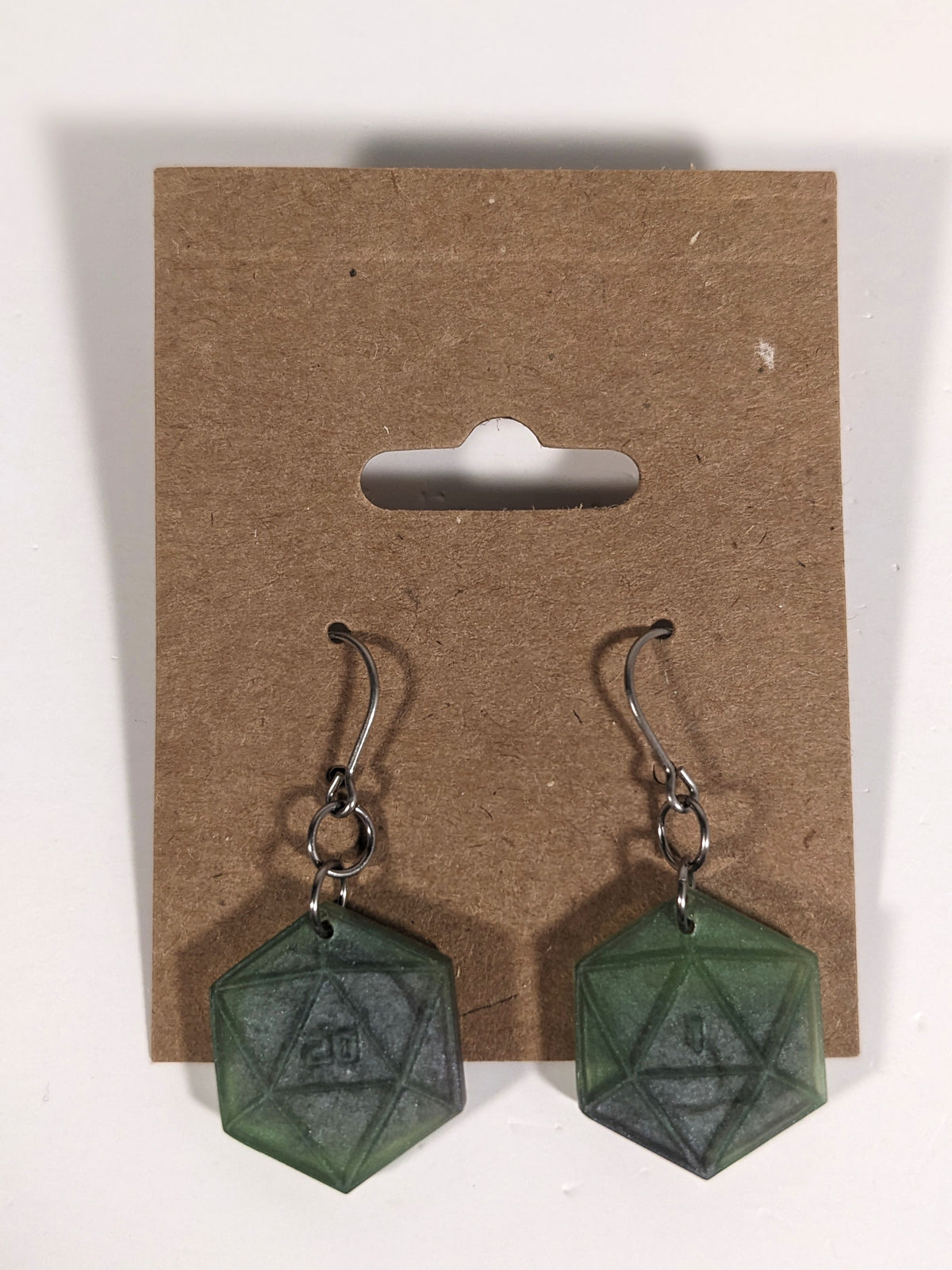 A pair of dark green and dark silver dangly, flat d20 earrings with silver color titanium and niobium hardware on a kraft earring card on a white background. The earring on the left has a 20 on it and the earring on the right has a 1 on it, both in a sci-fi style font.