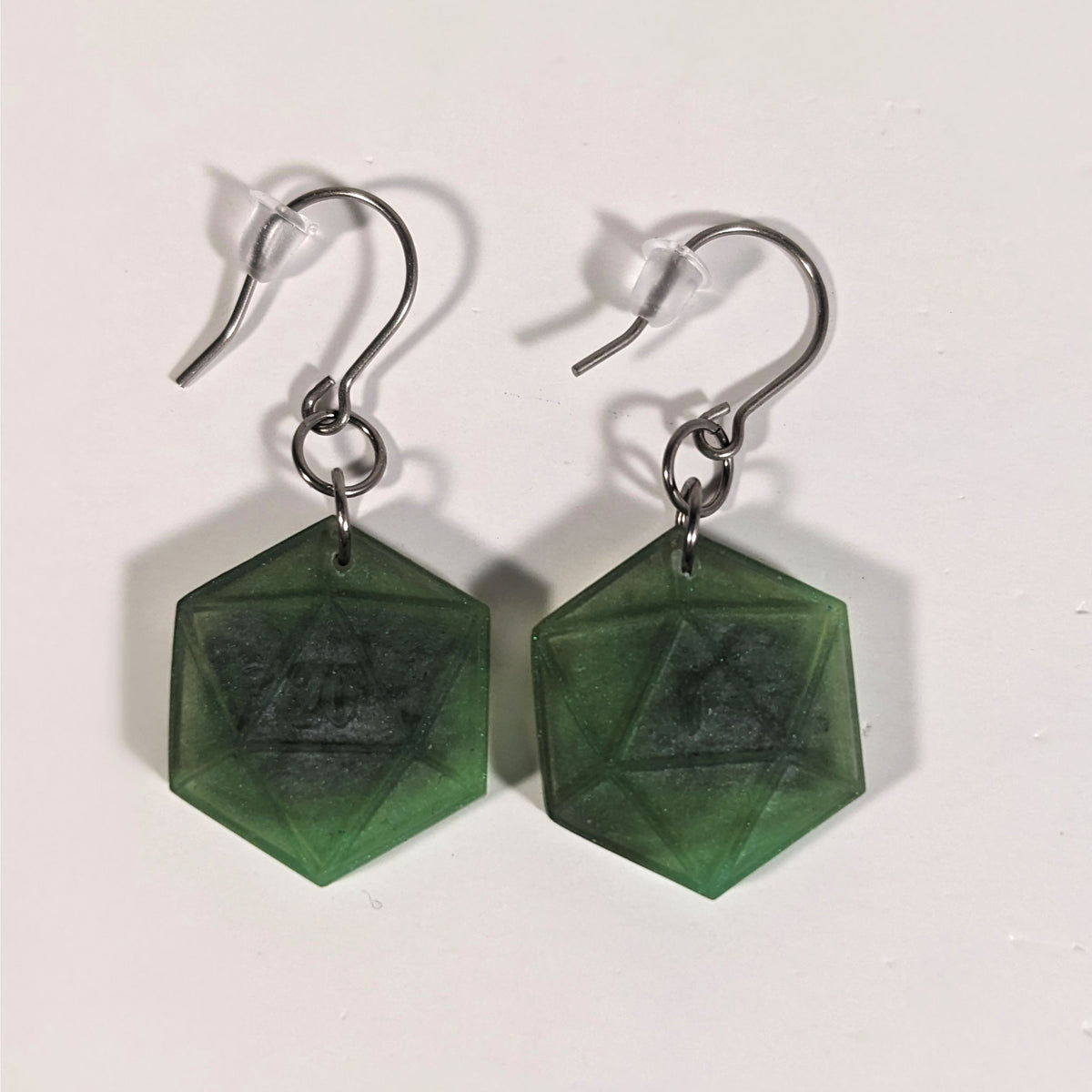 A pair of dark green and dark silver dangly, flat d20 earrings with silver color titanium and niobium hardware and bell shaped rubber backs on a white background. The earring on the left has a 20 on it and the earring on the right has a 1 on it, both in a fantasy style font.