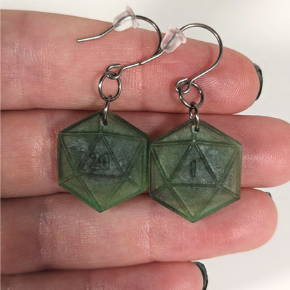 A pair of dark green and dark silver dangly, flat d20 earrings with silver color titanium and niobium hardware with bell shaped rubber backs held on on a hand on a white background. The earring on the left has a 20 on it and the earring on the right has a 1 on it, both in a fantasy style font.