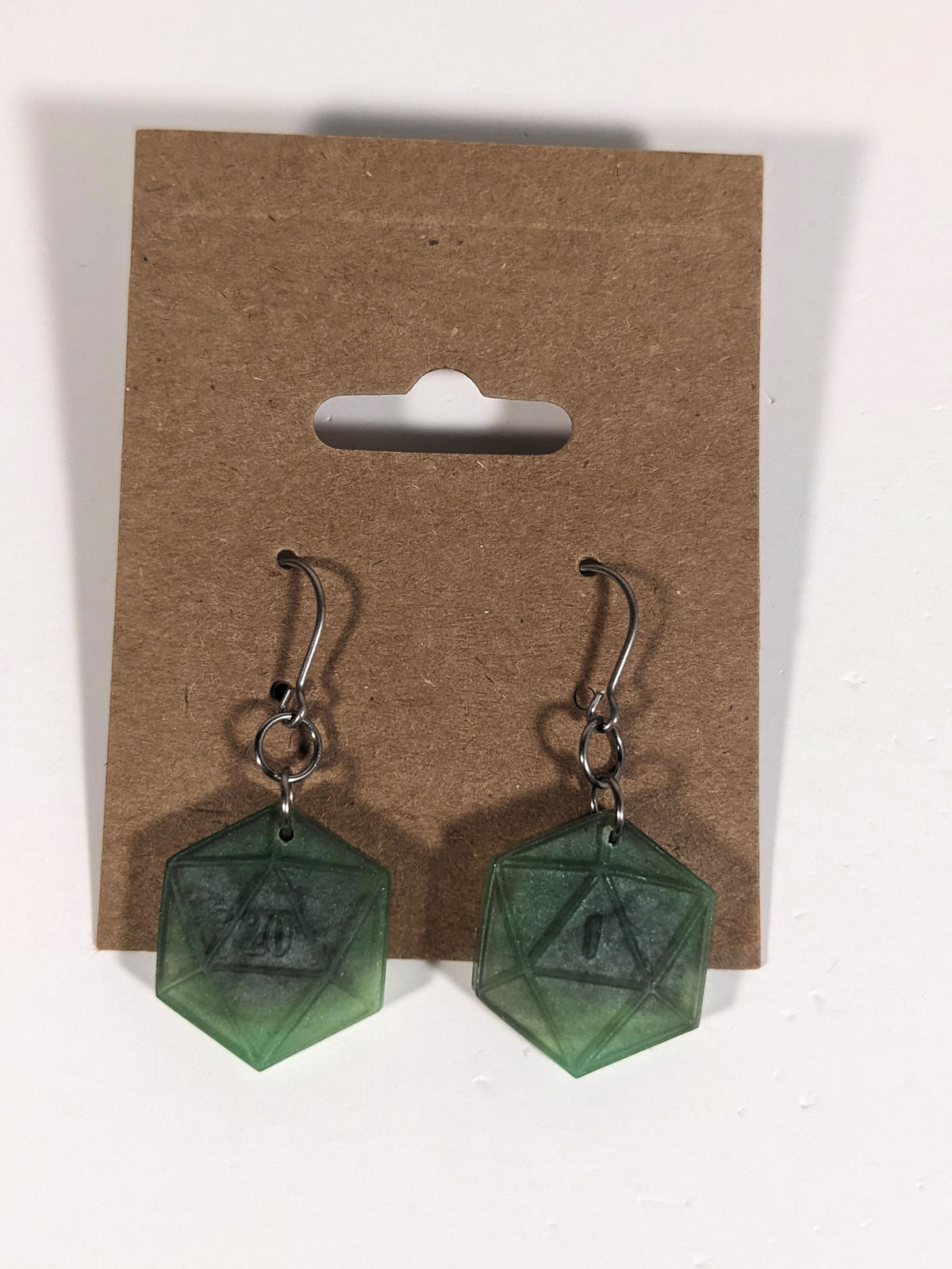 A pair of dark green and dark silver dangly, flat d20 earrings with silver color titanium and niobium hardware on a kraft earring card on a white background. The earring on the left has a 20 on it and the earring on the right has a 1 on it, both in a fantasy style font.