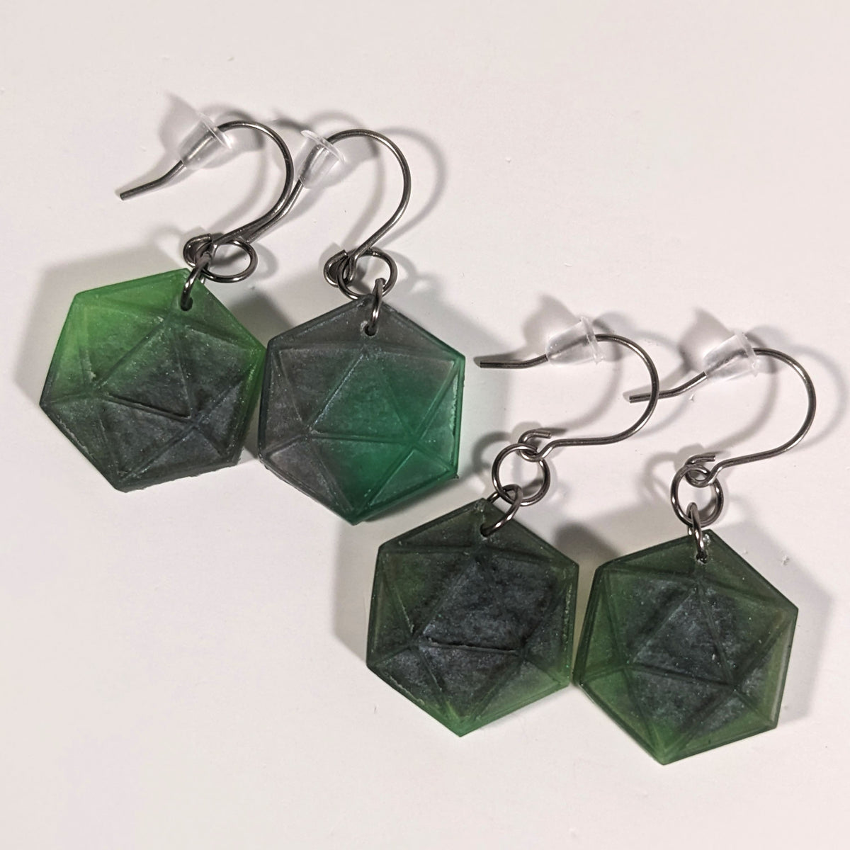 Two pairs of dark green and dark silver dangly, flat d20 earrings with silver color titanium and niobium hardware and bell shaped rubber backs on a white background.