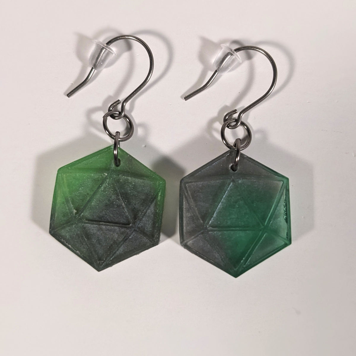 A pair of dark green and dark silver dangly, flat d20 earrings with silver color titanium and niobium hardware and bell shaped rubber backs on a white background.