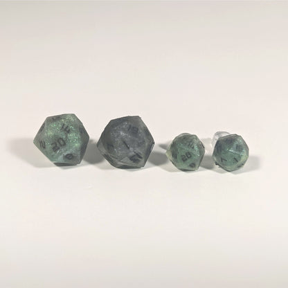Two pairs of handmade d20 earrings with titanium hardware that are green and dark silver with black numbers on a white background sit in a line with the larger pair on the left and the smaller pair on the right. The earrings on the left of each pair centers the 20 face and the earrings on the right of each pair centers the 1 face.