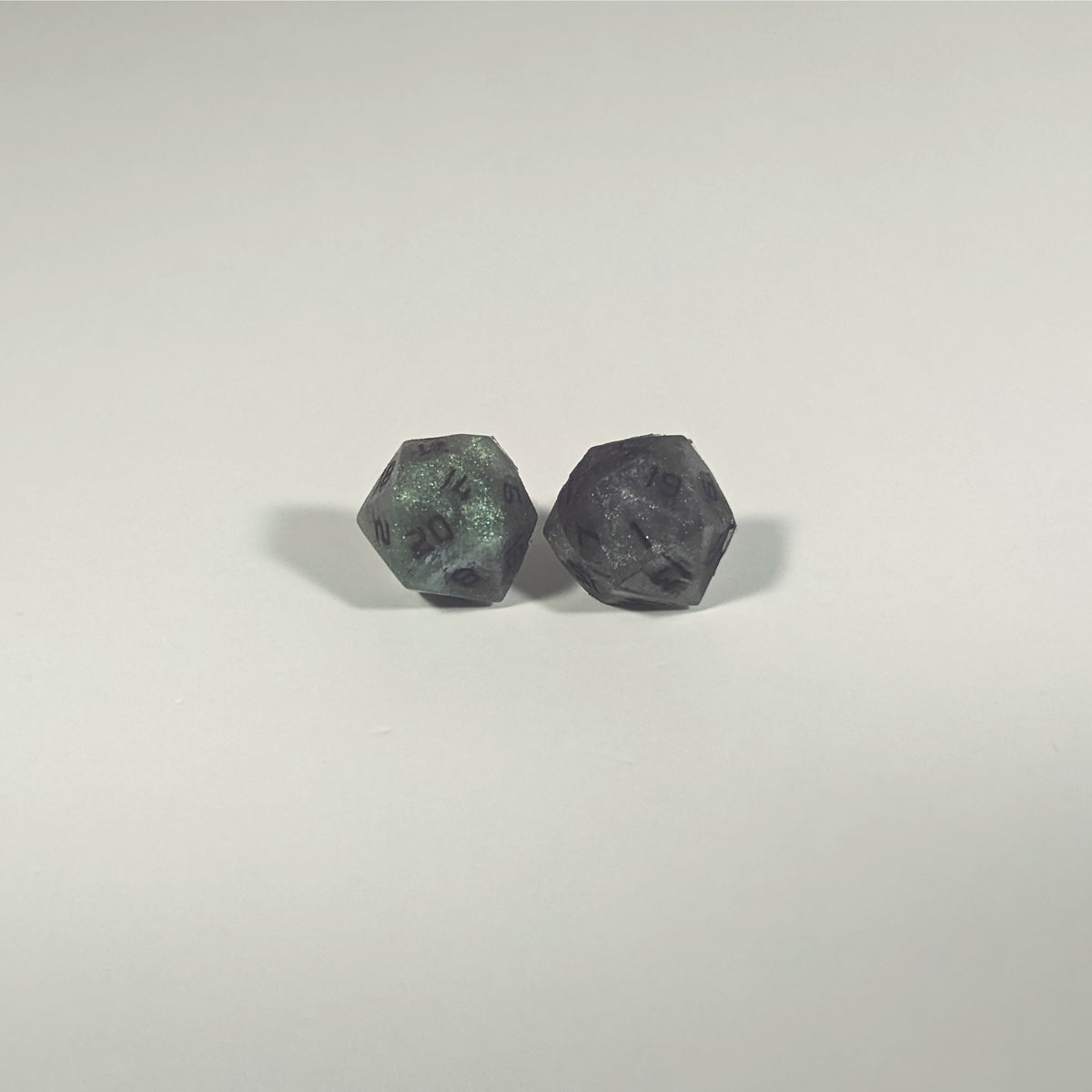 A single pair of the larger size handmade d20 earrings with titanium hardware that are green and dark silver with black numbers on a white background. The earring on the left centers the 20 face and the earring on the right centers the 1 face.