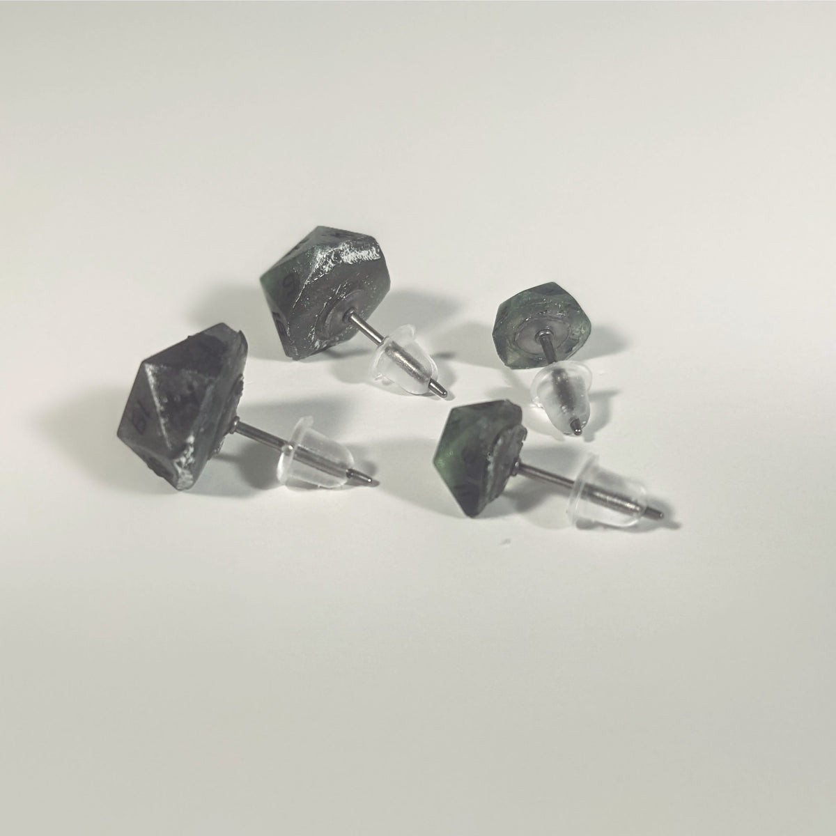 Two pairs of handmade d20 earrings that are green and dark silver with black numbers turned to show the titanium hardware and bell shaped rubber backs.