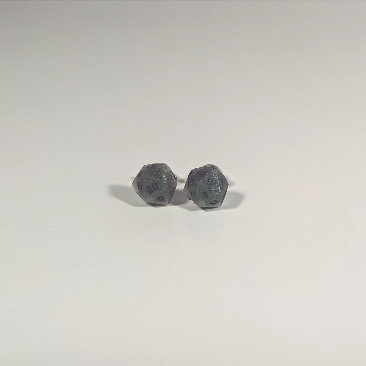 A single pair of the smaller size handmade d20 earrings with titanium hardware that are green and dark silver with black numbers on a white background. The earring on the left centers the 20 face and the earring on the right centers the 1 face.