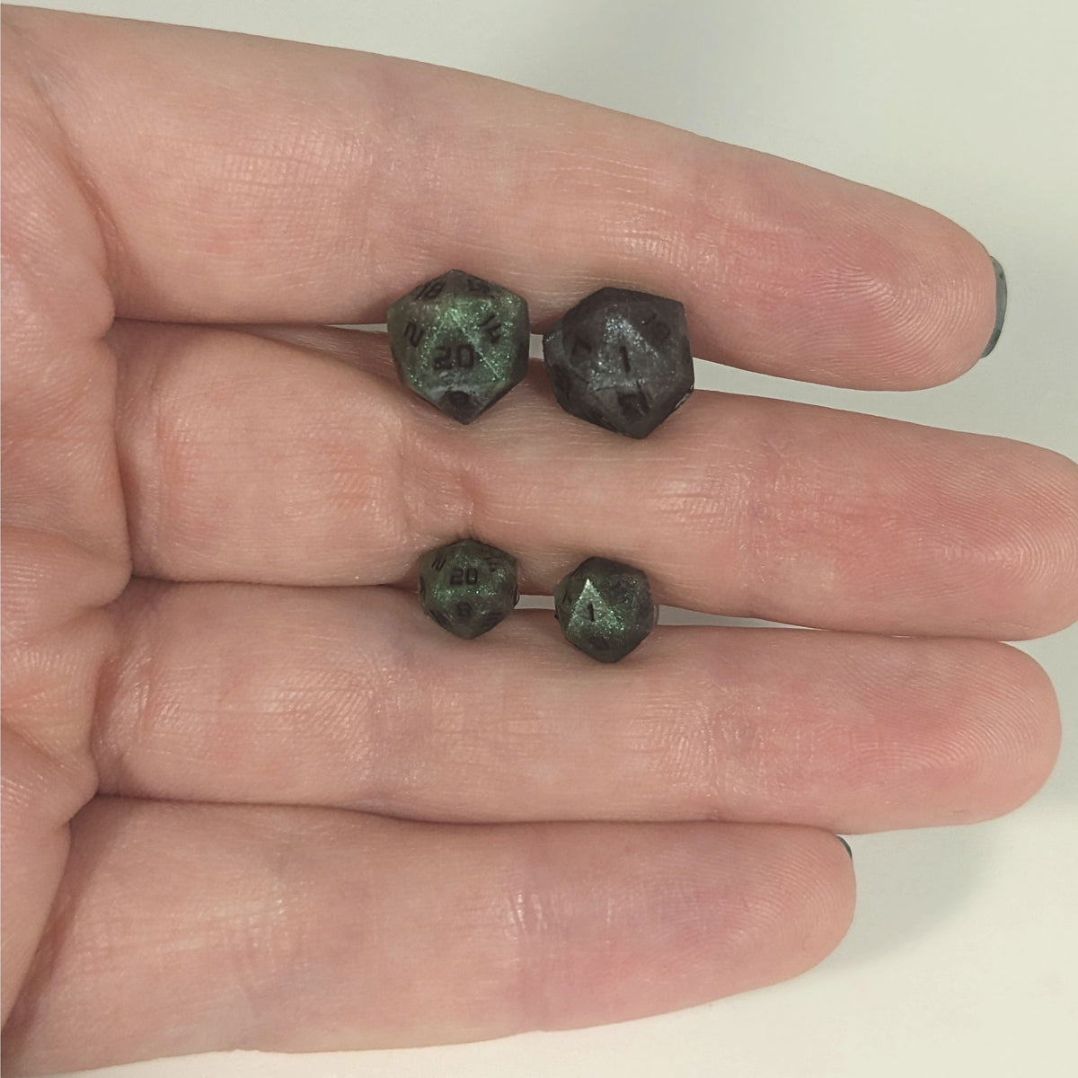 Two pairs of handmade d20 earrings with titanium hardware that are green and dark silver with black numbers held between the fingers of a flat hand. The larger pair is between the pointer and middle finger, the smaller between the middle and ring finger. The earrings on the left of each pair centers the 20 face and the earrings on the right of each pair centers the 1 face.