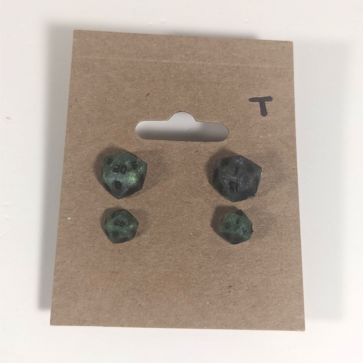 Two pairs of handmade d20 earrings with titanium hardware that are green and dark silver with black numbers on a kraft brown earring card with a T in the top right corner. The earrings on the left of each pair centers the 20 face and the earrings on the right of each pair centers the 1 face.