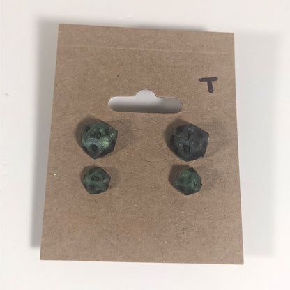 Two pairs of handmade d20 earrings with titanium hardware that are green and dark silver with black numbers on a kraft brown earring card with a T in the top right corner. The earrings on the left of each pair centers the 20 face and the earrings on the right of each pair centers the 1 face.