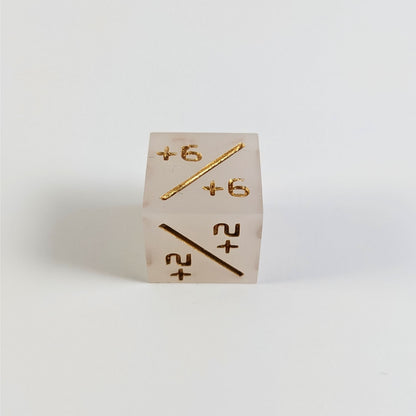 A low angle photo of a white positive counter d6 with gold numbers sits on a white background with the +6/+6 face up and the +2/+2 face pointed towards the camera.