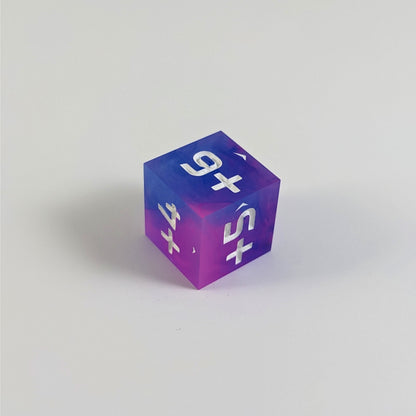 A mid angle photo of a pink and blue positive spindown d6 with white numbers sits on a white background with the corner of the +6, +5, and +4 faces angled toward the camera.