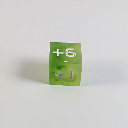 A low angle photo of a yellow green positive spindown d6 with white numbers sits on a white background with the +6 face up and the +1 face pointed towards the camera.