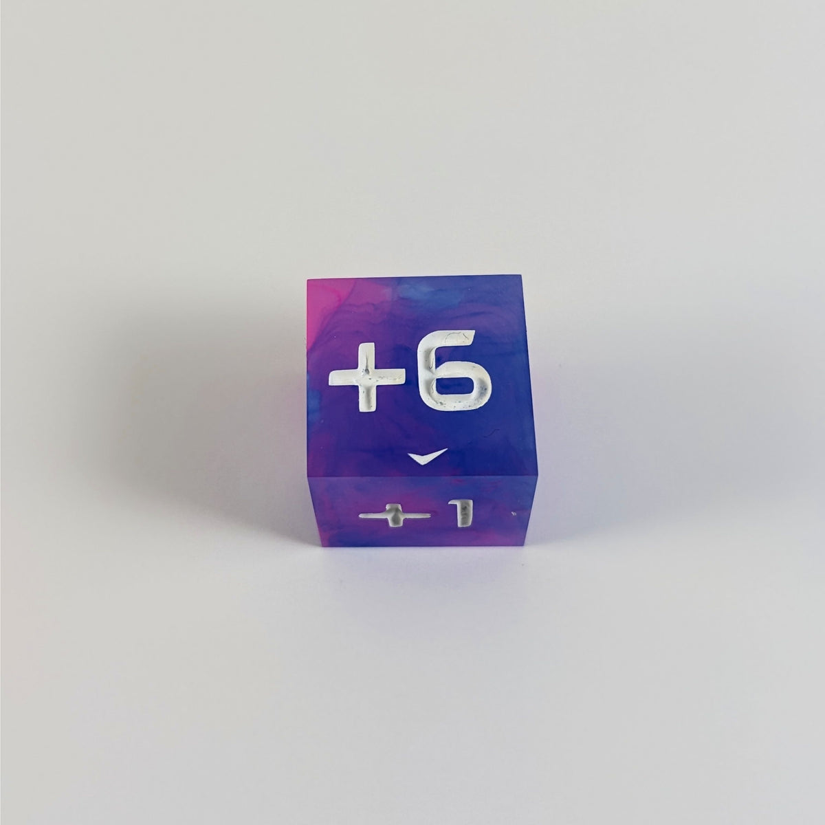 A high angle photo of a pink and blue positive spindown d6 with white numbers sits on a white background with the +6 face up.