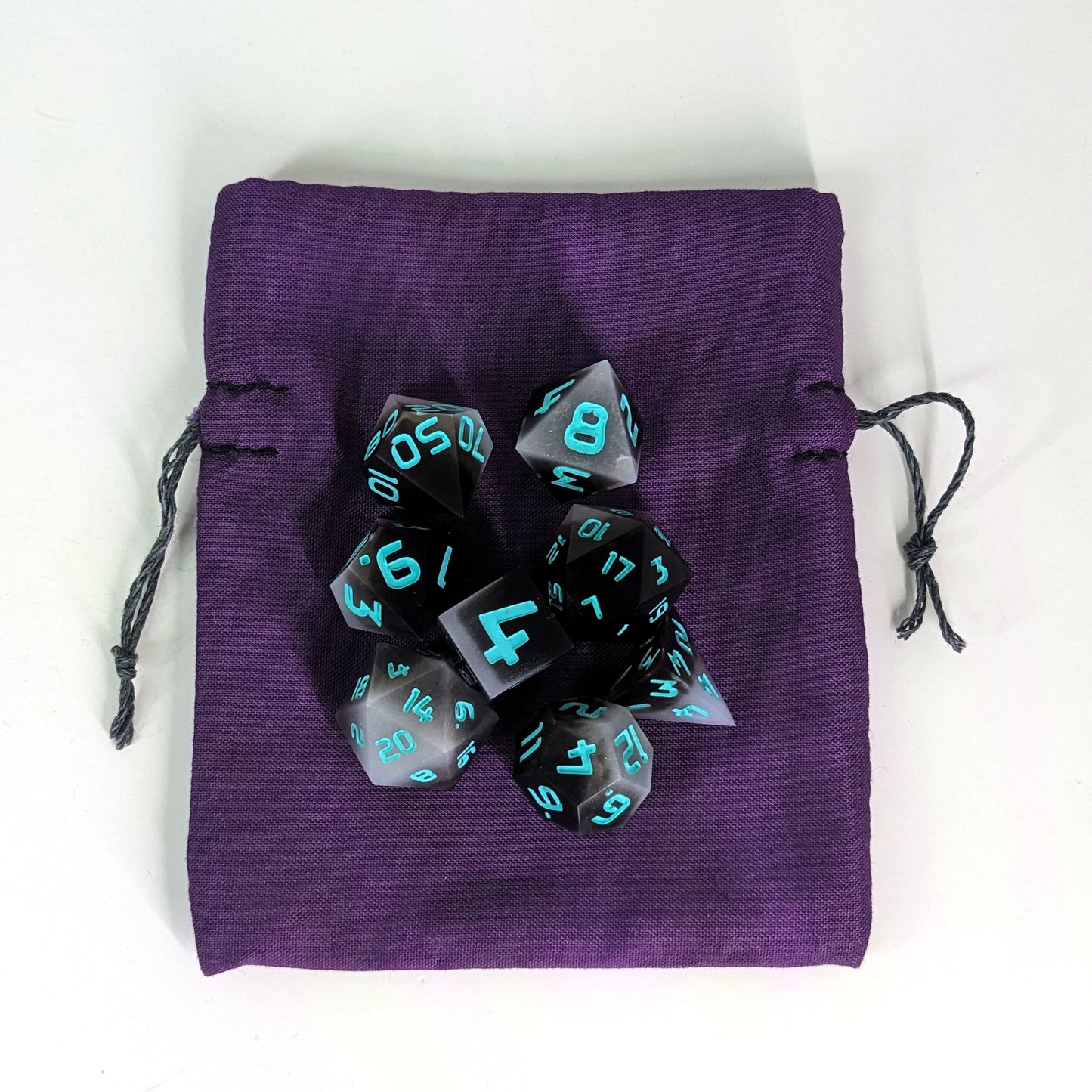 A small, handmade dice bag that is purple lays flat on a white background with a set of brown and white dice with teal numbers sitting on top of it.